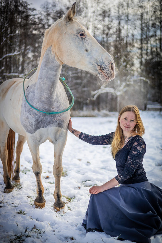 Frau im blauen Kleid kniet neben einem weißen Pferd bei einem winterlichen Pferdeshooting im Schnee.