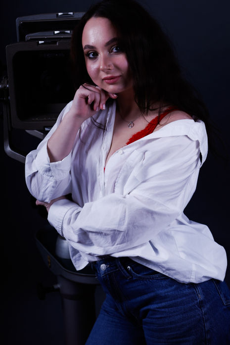 Frau mit Brille in weißer Bluse posiert mit einer Hand am Gesicht bei einem Studio-Portrait.