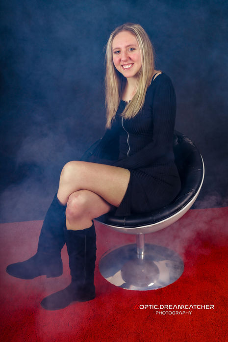 Frau in schwarzem Kleid sitzt auf einem Hocker vor einem dunklen Hintergrund mit rotem Boden.