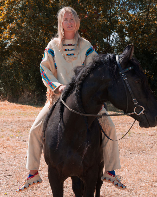 Blonde Frau in hellem Fransen-Outfit reitet auf einem schwarzen Pferd bei einem sommerlichen Reitportrait.