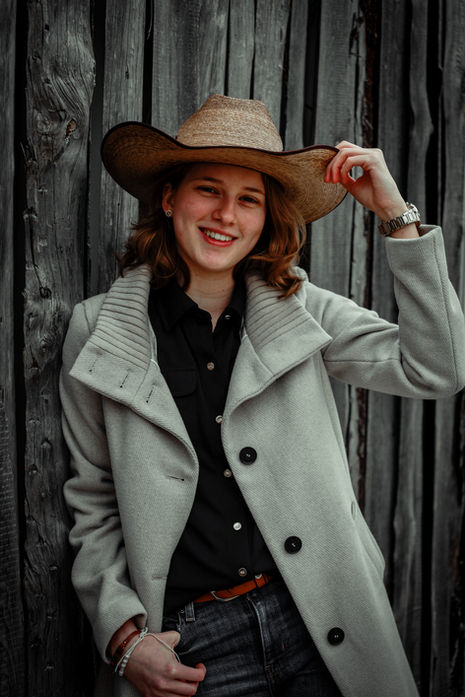 Rothaarige Frau mit Cowboyhut und hellem Mantel posiert vor einer rustikalen Holzkulisse.