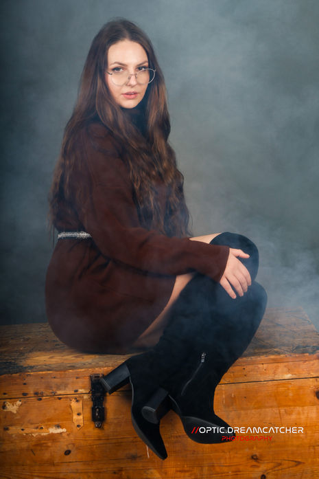 Frau in dunklem Outfit sitzt auf einer Holzkiste bei einem stimmungsvollen Studio-Portrait mit Nebel.