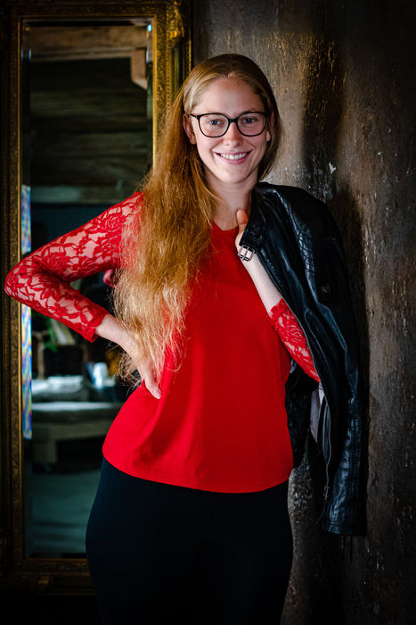 Frau mit Brille in rotem Oberteil und schwarzer Lederjacke posiert in einer dunklen Indoor-Location.
