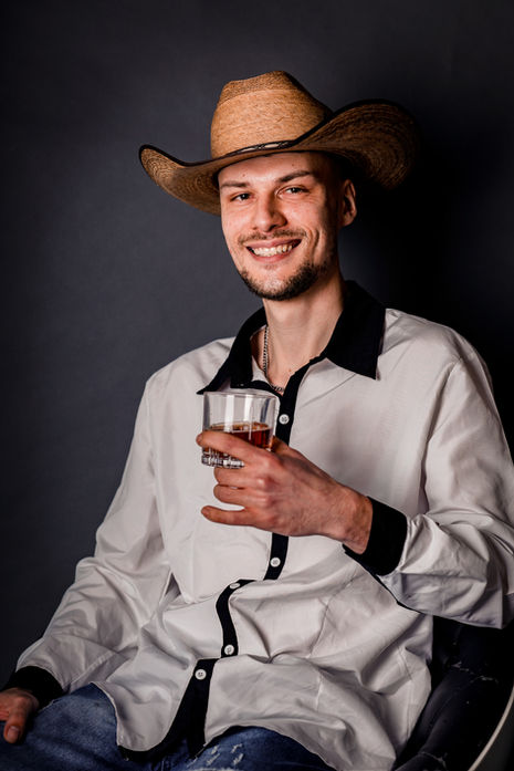 Lächelnder Mann mit Cowboyhut und hellem Hemd sitzt bei einem lockeren Country-Portrait.