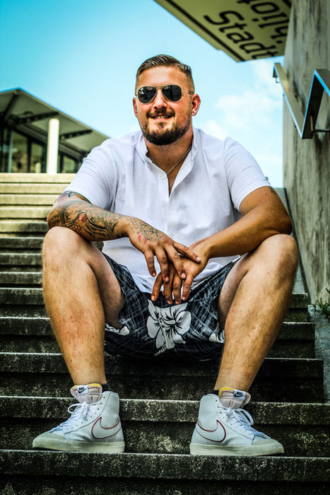 Mann mit Sonnenbrille, weißem Poloshirt und Shorts sitzt auf einer Treppe bei einem urbanen Outdoor-Shooting.