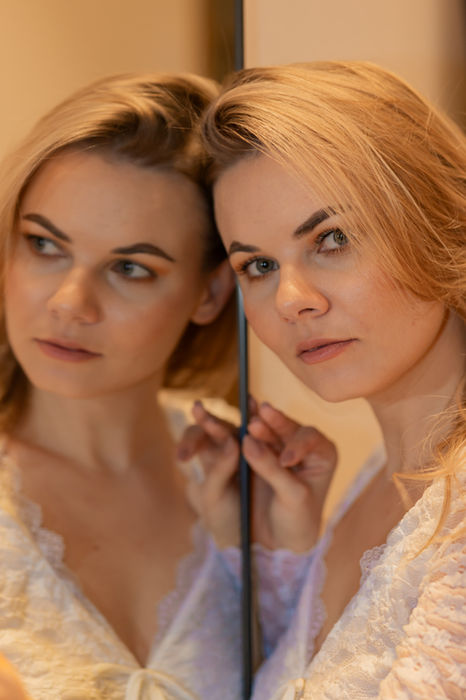 Nahaufnahme einer Frau in weißer Spitze neben einem Spiegel, der ihr Gesicht weich reflektiert.