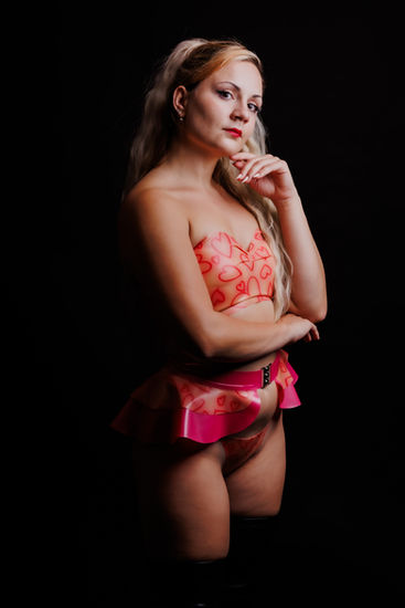 Frau in roter Lingerie steht vor schwarzem Hintergrund und posiert mit einer Hand am Gesicht.