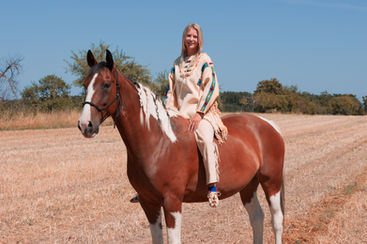 Blonde Frau sitzt auf einem braun-weißen Pferd bei einem Pferdeshooting auf einem trockenen Feld.