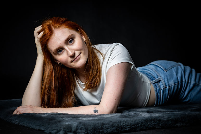 Rothaarige Frau in weißem Shirt und Jeans bei einem entspannten Portraitshooting vor dunklem Hintergrund.