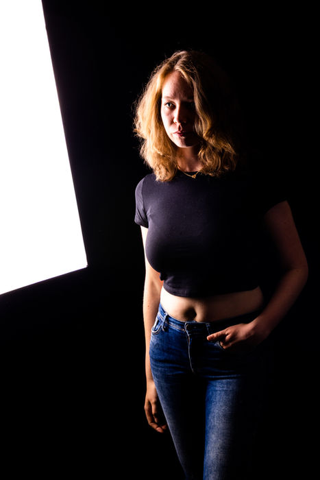 Frau in schwarzem Oberteil und Jeans steht im Seitenlicht bei einem kontrastreichen Studio-Portrait.