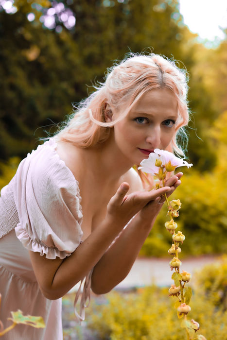 Blonde Frau in hellem Kleid beugt sich über eine Blüte bei einem verträumten Outdoor-Portrait im Grünen.