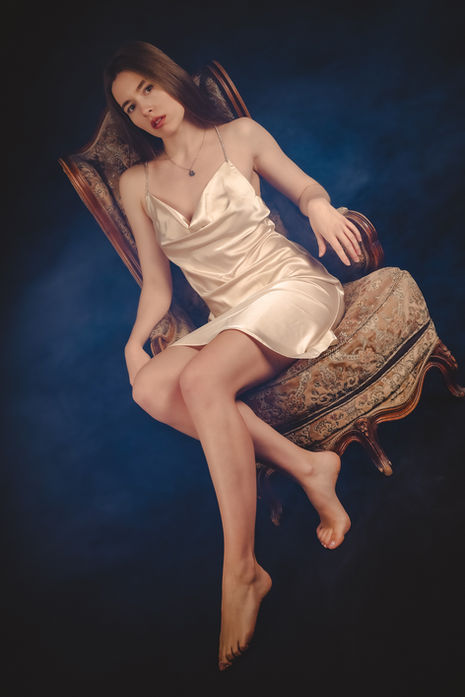 Frau in hellem Outfit liegt entspannt auf einem Vintage-Sessel vor dunklem Studiohintergrund.