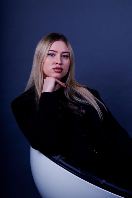 Blonde Frau in schwarzem Outfit sitzt auf einem modernen Stuhl und stützt das Kinn auf eine Hand.