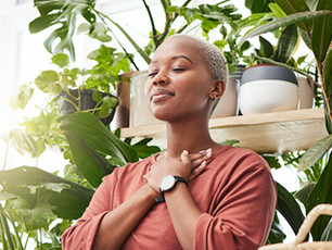 A person in a pink shirt stands among lush green plants, eyes closed, hands on chest, exuding calm and contentment. Bright light filters in.
