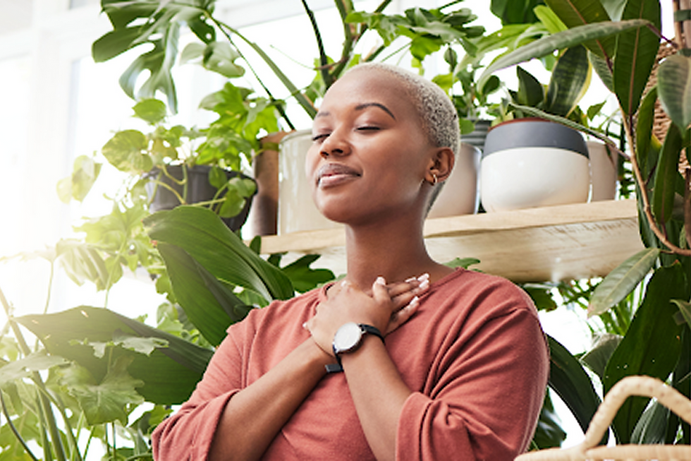 A person in a pink shirt stands among lush green plants, eyes closed, hands on chest, exuding calm and contentment. Bright light filters in.