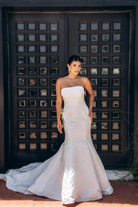 Modern Strapless Fit-and-Flare Wedding Dress with Mermaid Skirt
For the bride seeking luxe elegance with a contemporary twist, Style 8011 is a modern strapless wedding dress that makes a striking statement down the aisle. Its fit-and-flare design hugs every curve in all the right places, accentuating the natural figure. A sleek and straight neckline with a subtle banded detail introduces the fitted strapless bodice also available with in unique textured fabric throughout elevates the gown’s clean and chic aesthetic into a fashion-forward design piece. In the back, this chic wedding gown is adorned with fabric-covered buttons from the banded top of the bodice to the hem of the dramatic flared mermaid skirt for a final bold statement in this modern fit-and-flare wedding dress. This wedding gown is also available in our plus range.