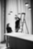 sw foto, eine person steht leicht erhöt auf einer leiter und bespricht sich mit einer weiteren. im bild ist ein tresen davor, und hinter ihnen ein fenster hinter einer gardien. edles, reduziertes reportage foto