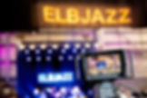 unscharf im hintergrund das elbjazz logo beleuchtet, sowie darunter eine bühne. im vordergrund das display einer kamera die gerade das konzert aufnimmt