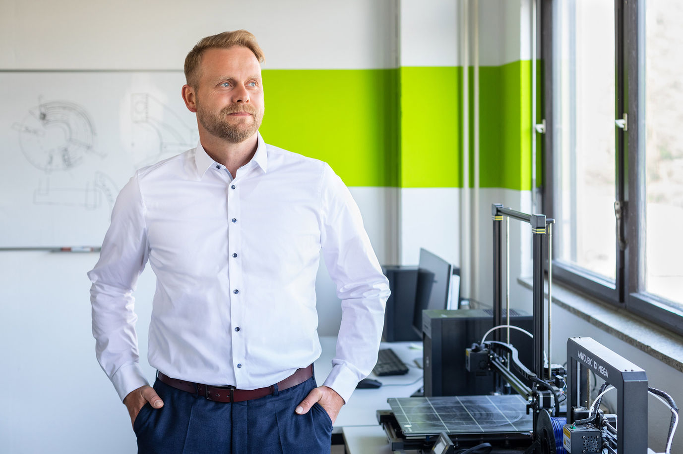 Businessportrait eines Mannes im weißen Hemd, stehend in einem hellen Büro neben einem 3D-Drucker, mit Blick zum Fenster und technischer Arbeitsumgebung im Hintergrund.