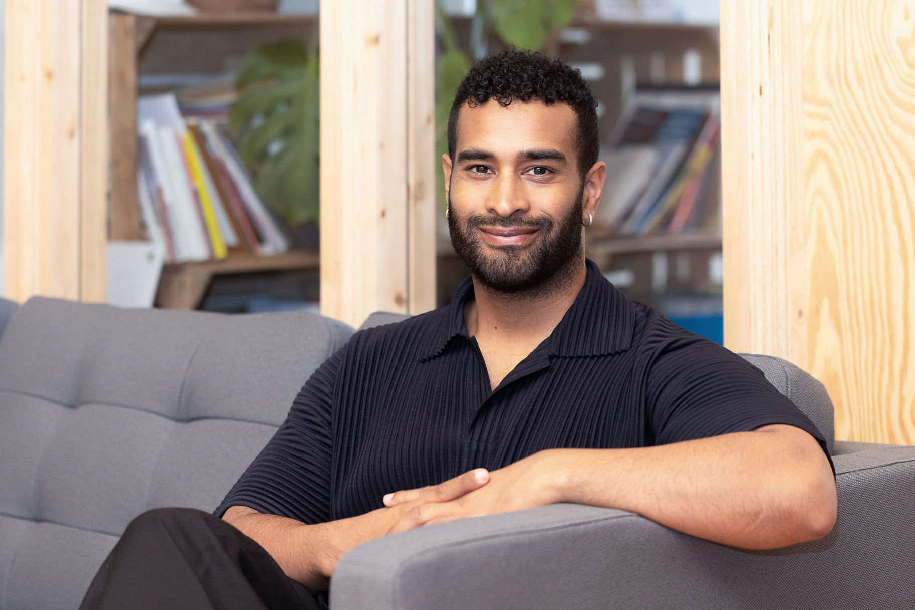 Portrait eines lächelnden Mannes mit Bart und kurzem lockigem Haar, sitzend auf einem grauen Sofa vor einer hellen Holzwand mit Bücherregal im Hintergrund in Berlin.