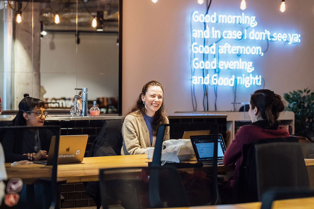 Drei Frauen im Gespräch an einem großen Arbeitstisch mit Laptops, warmem Licht und blau leuchtendem Schriftzug im Hintergrund – Businessfotografie in Berlin.