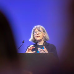 Rednerin auf der Angel Conference im Humboldt Forum Berlin, gerahmt zwischen zwei unscharfen Silhouetten im Publikum vor violettem Bühnenlicht – atmosphärische Eventfotografie und Konferenzfotografie in Berlin mit cineastischer Komposition und Fokus auf Vortragssituation und Publikumsperspektive.
