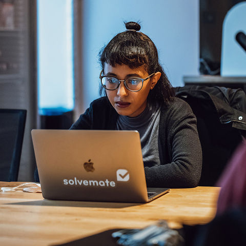 Eine Mitarbeiterin sitzt an einem Holztisch vor einem Laptop und arbeitet konzentriert in einer modernen Büroumgebung. Im Vordergrund ist unscharf eine weitere Person zu sehen, im Hintergrund das Logo des Unternehmens.