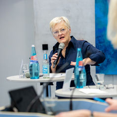 Eine bekannte Politikerin, Renate Künast, spricht bei einem Event in ein Mikrofon. Nur ihr Gesicht und das Mikrofon sind scharf, während der Vorder- und Hintergrund stark unscharf und überwiegend Blau sind.