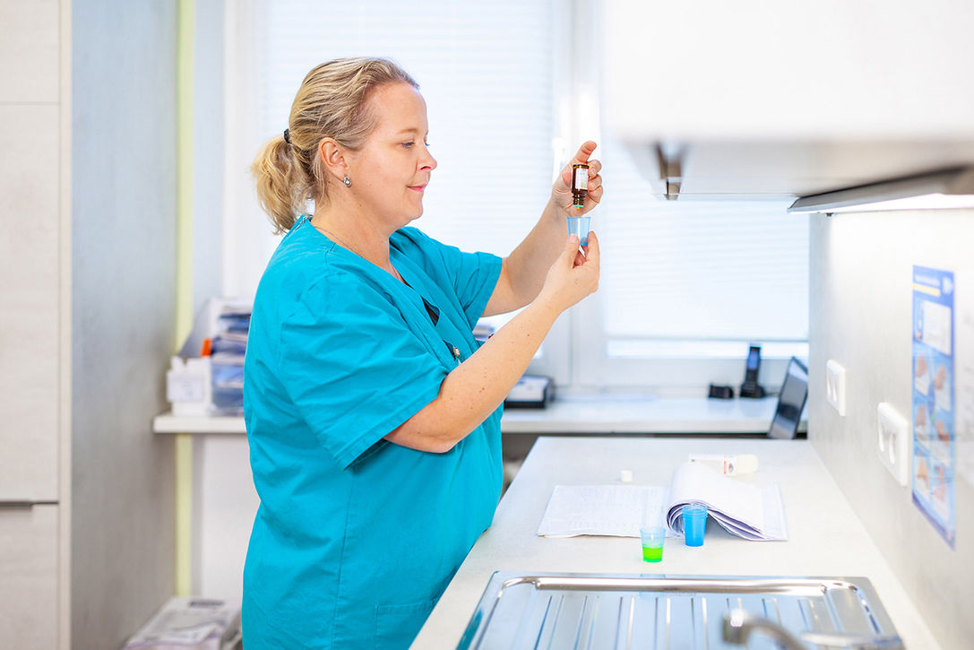 Corporate Fotografie im Gesundheitswesen: Schwester im blauen Kittel bereitet im Pflegeraum Medikamente vor, tropft Flüssigkeit in kleinen Plastikbecher, davor auf der Theke eine Patientenakte und weitere Becher.