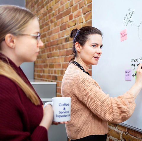 Eine Frau erklärt an einer Whiteboard-Zeichnung, die mit Notizen versehen ist, während zwei Kolleginnen aufmerksam zuhören. Die Backsteinwand im Hintergrund verleiht dem Raum einen kreativen Charakter.