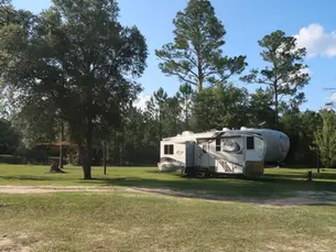 RV camper parked at a shaded campsite in Escambia County, Florida near the Blackwater River, surrounded by pine trees and open grassy camping area.