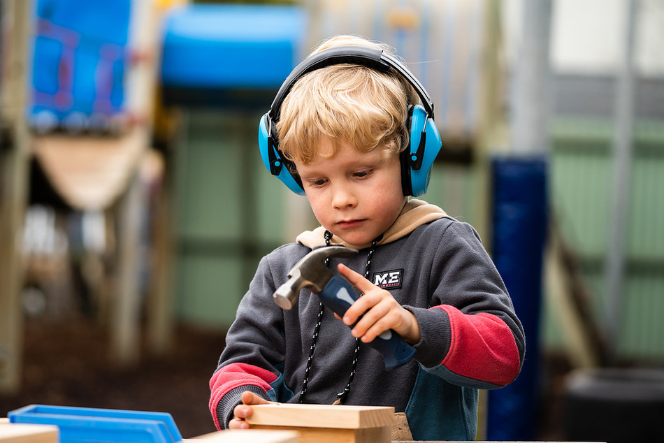 Boy wearing headphones while building something with wood and hammer and nails at Kindy in Halswell