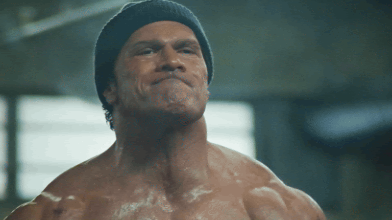 Dwayne Johnson, in prosthetics and fight gear, sits exhausted in a locker room surrounded by shadows in The Smashing Machine (2025), directed by Benny Safdie.