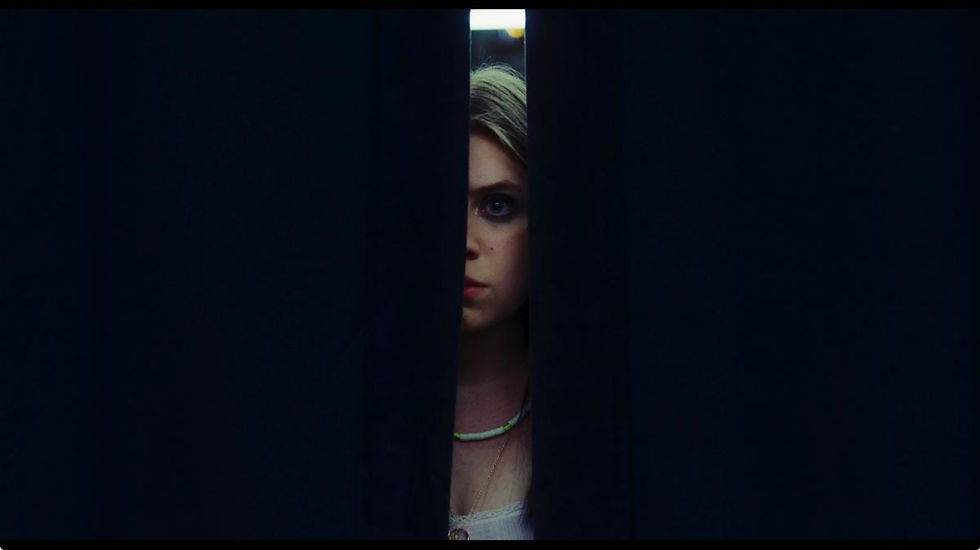 Kate Hallett peering through curtains in a dim room, watching someone off-screen in the first-look image from Sweetness.