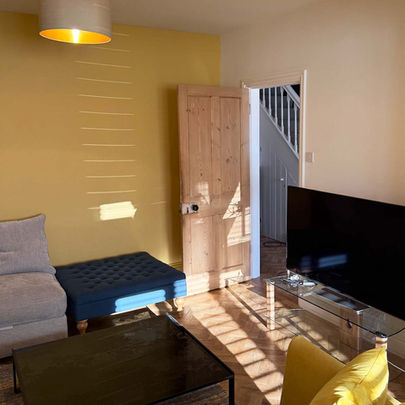 Professionally painted lounge with a mustard yellow feature wall, warm off-white walls and matching ceiling, wooden floor, coordinating furniture, and an open stripped-pine vintage door.