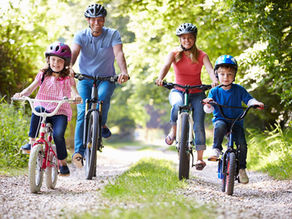 Family cycling on a sunny path, surrounded by green trees. Two kids lead on small bikes, followed by adults on larger bikes, all smiling.