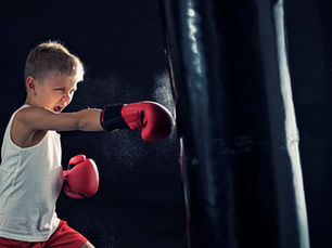 Young boy in a white tank top and red gloves punches a black punching bag with determination in a dark setting.