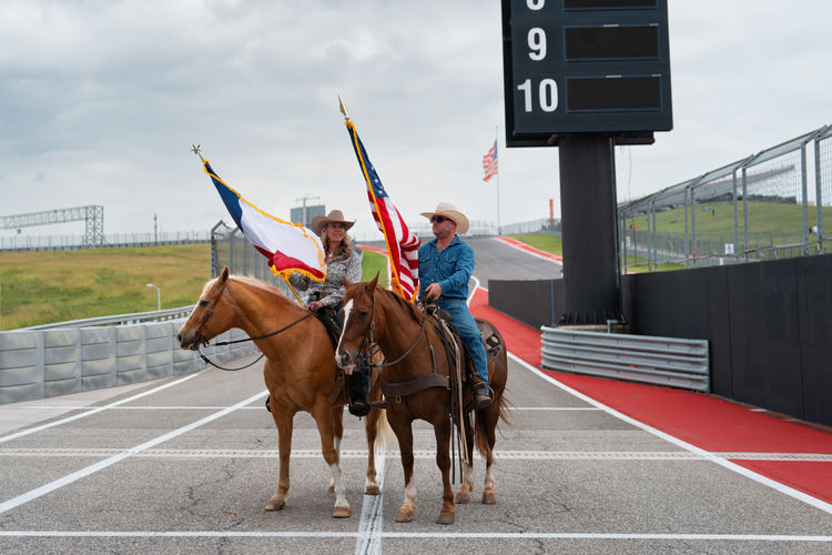 2 riders hold flags on horseback at pit exit of Circuit of the Americas, 2025 GT World Challenge America