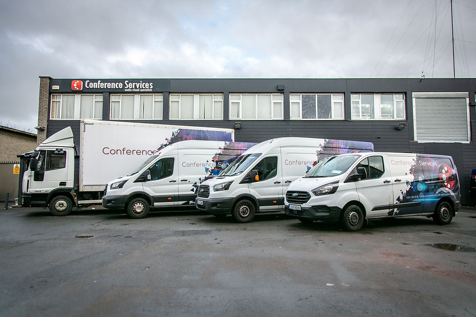 A fleet of various white vans and trucks are parked in a row outside a large, modern building with a grey facade. The vehicles suggest transportation and logistical support for event management.