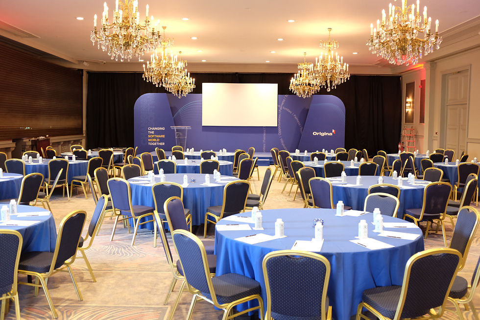 An elegant event venue is shown with round tables draped in blue tablecloths and white chairs. The stage features a screen and subtle lighting. The focus is on a sophisticated interior, suggesting a sustainable approach to event design with reusable and low-waste elements.