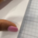 Close-up of a hand with pink polished nail holding white paper over graph paper, with a metal edge along the side.