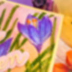 Purple crocus on pastel card with "en" text, surrounded by green stems and soft colors. Bright, cheerful floral design.