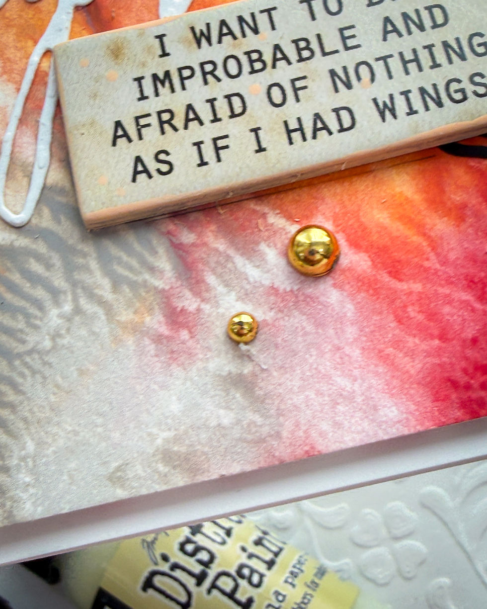 Text on a colorful textured background reads "I WANT TO DO IMPROBABLE AND AFRAID OF NOTHING AS IF I HAD WINGS." Gold buttons and paint tube visible.