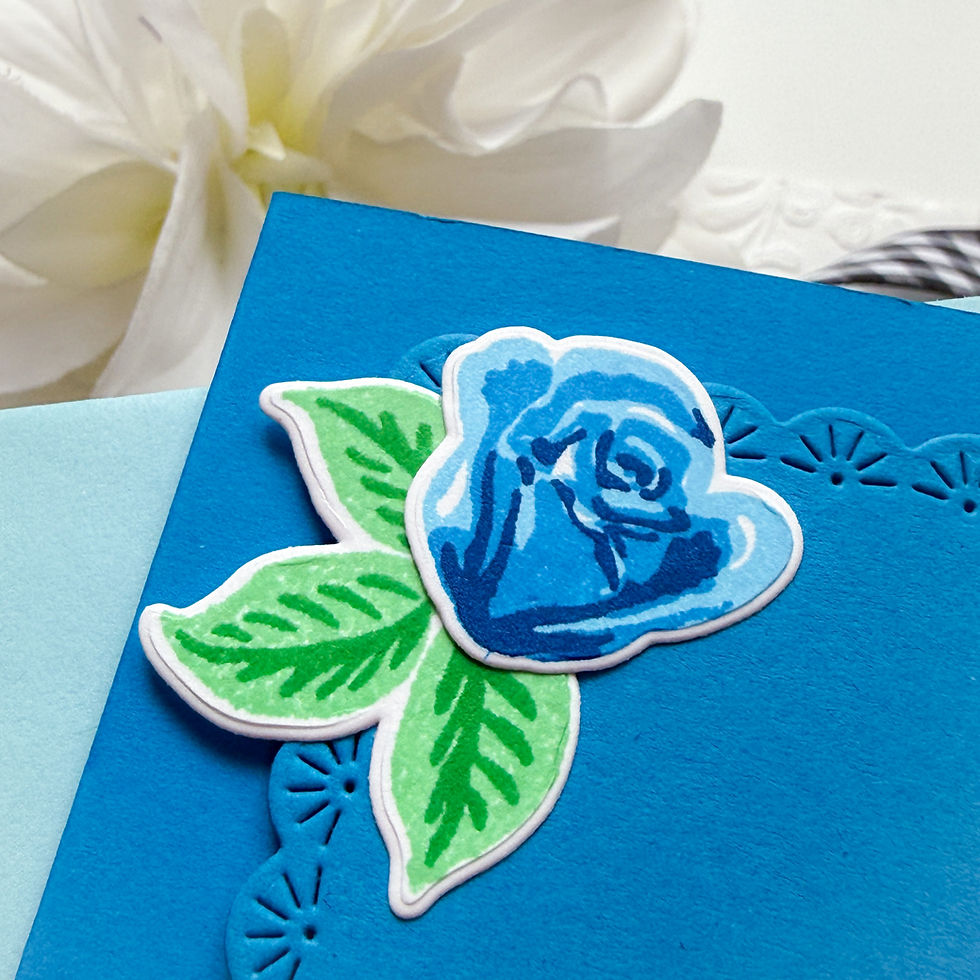 Handmade blue card with a blue rose and green leaves on textured paper, set against a soft white flower background.