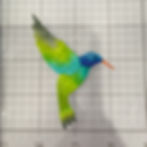 Colorful hummingbird ornament with blue head, green and turquoise wings, and orange beak on a gridded cutting mat.