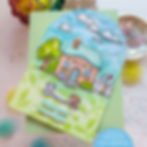 Pastel handmade card with a house, clouds, trees, two dogs, and text Wish you were here on a leafy blue-green background.