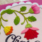 Embossed card with colorful flowers and green leaves, set against a pink background. Elegant text partially visible at the bottom. Mood is cheerful.