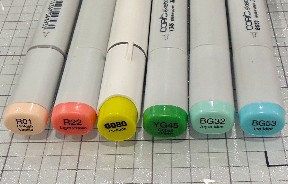Six Copic markers in various colors (pinkish vanilla, light prawn, limeade, cobalt green, aqua mint, ice mint) on a gridded surface.