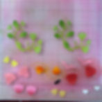 Colorful paper cutouts of leaves and flowers on a pink grid surface, featuring green branches and various petal shapes in yellows, pinks, and reds.