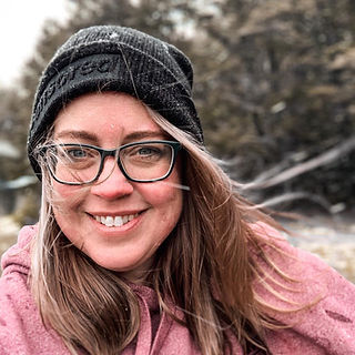 Blonde woman with glasses, pink sweater and black hat smiles in the snow